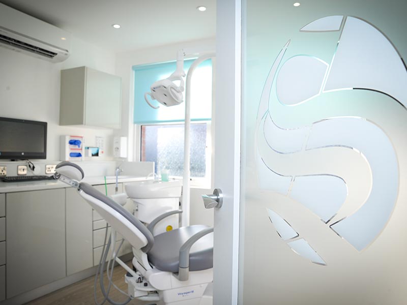 clean, bright treatment room with grey dental chair and sarum logo etched onto glass door
